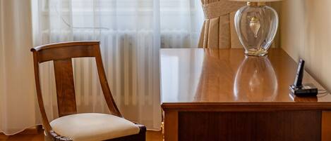 A wooden desk with a clear glass lamp, a wooden chair, a patterned rug, and a view through sheer curtains to a window.