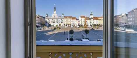 A view from a window of a building with a balcony, overlooking a square with historic buildings and a fountain.