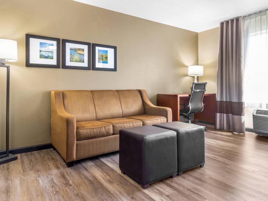 A hotel room with a brown leather sofa, two black ottomans, a standing lamp, a side table with a lamp, and framed pictures on the wall.