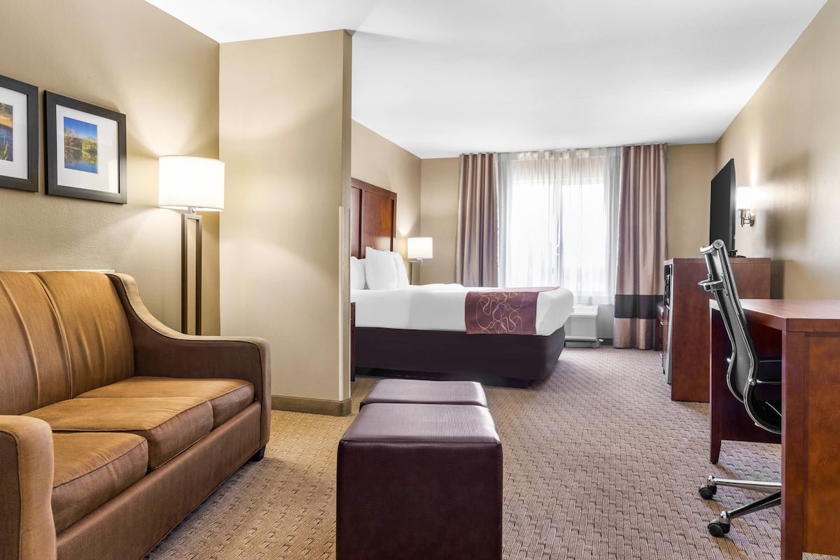 A hotel room with a brown leather sofa, a wooden desk, a bed with white linens, and a large window with curtains.