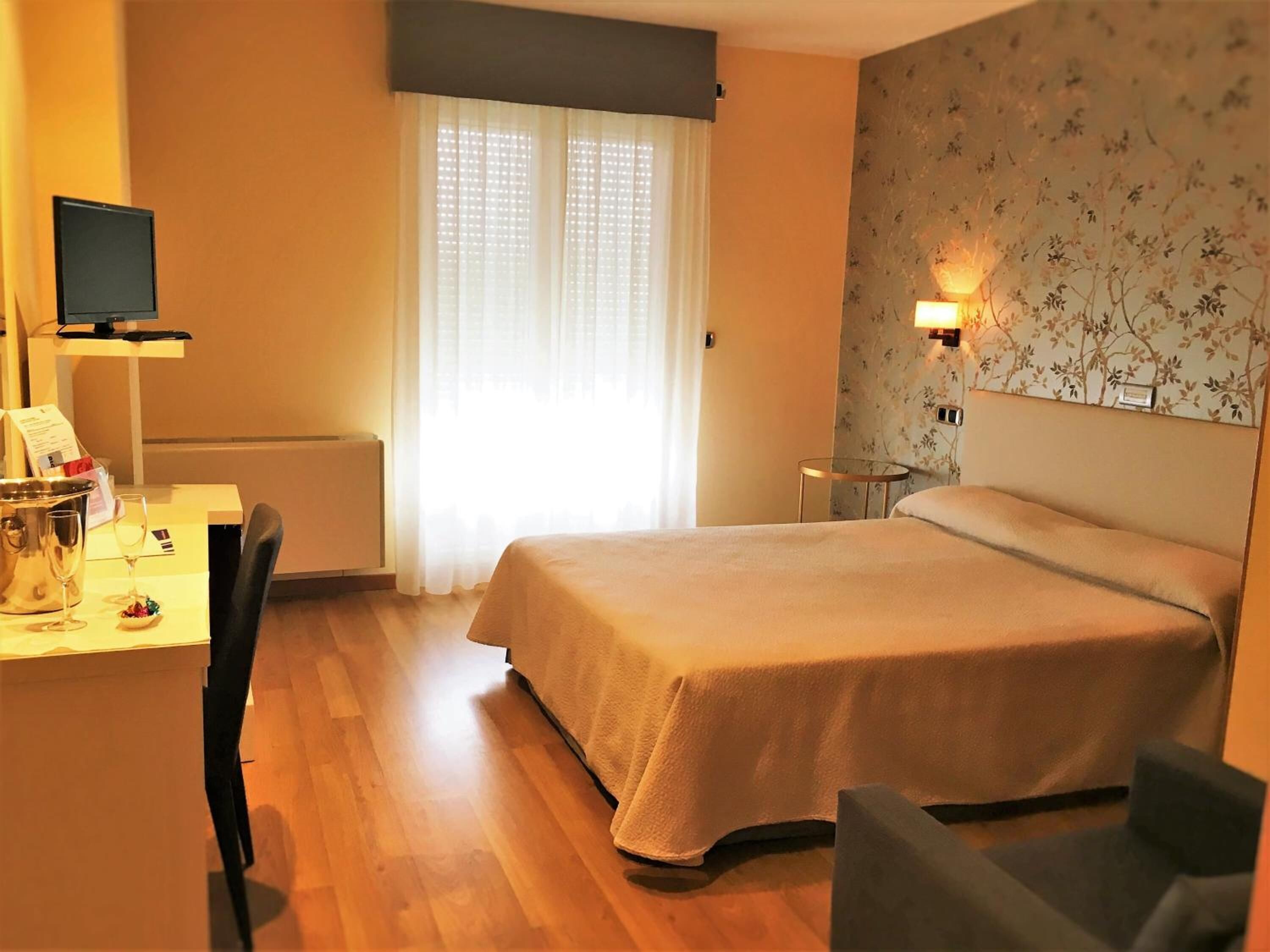 A hotel room with a bed, a desk with a computer, a chair, a small table, and a wall-mounted TV.