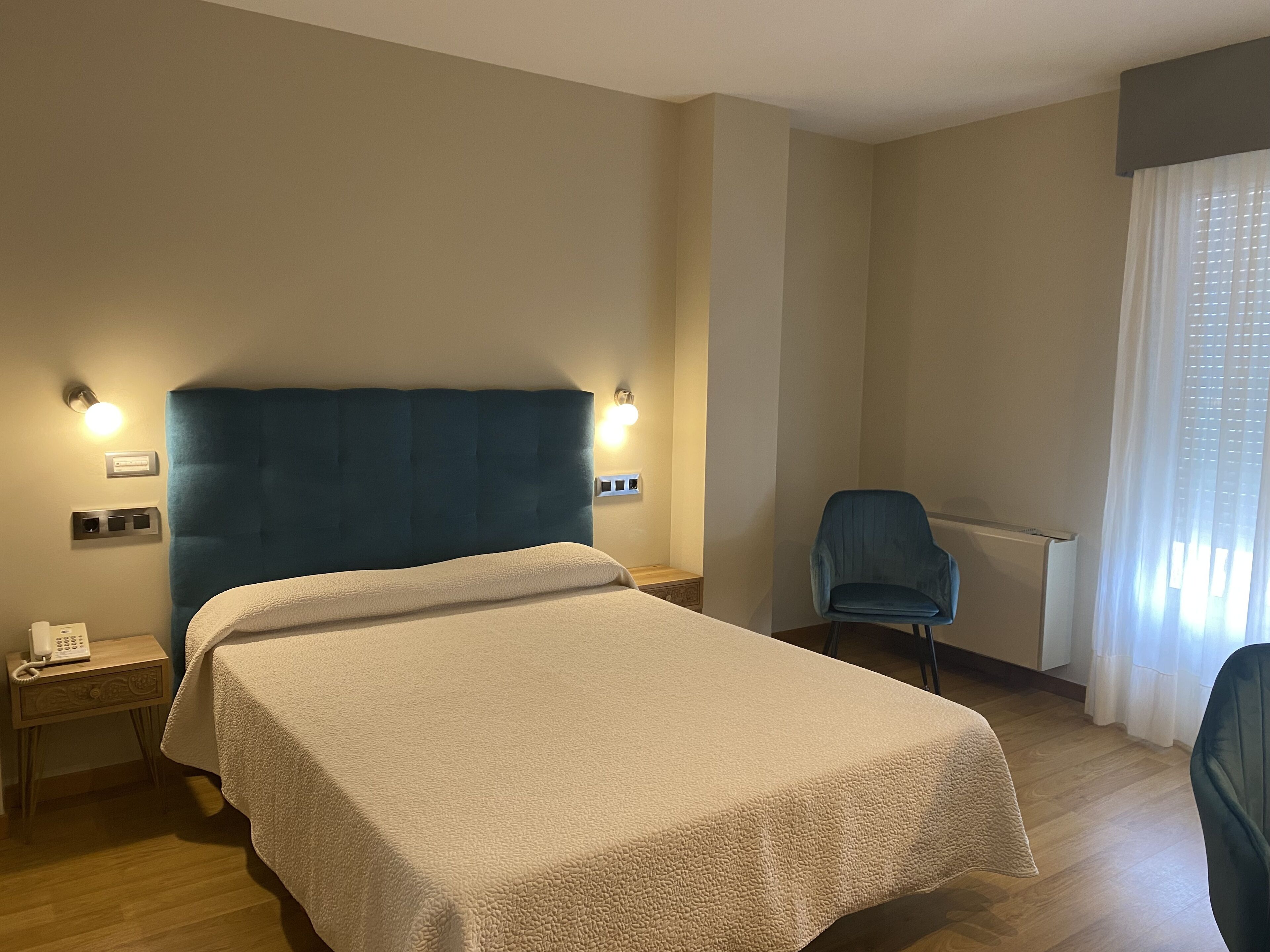 A hotel room with a large bed, two bedside lamps, a chair, a desk with a phone, and a window with curtains.