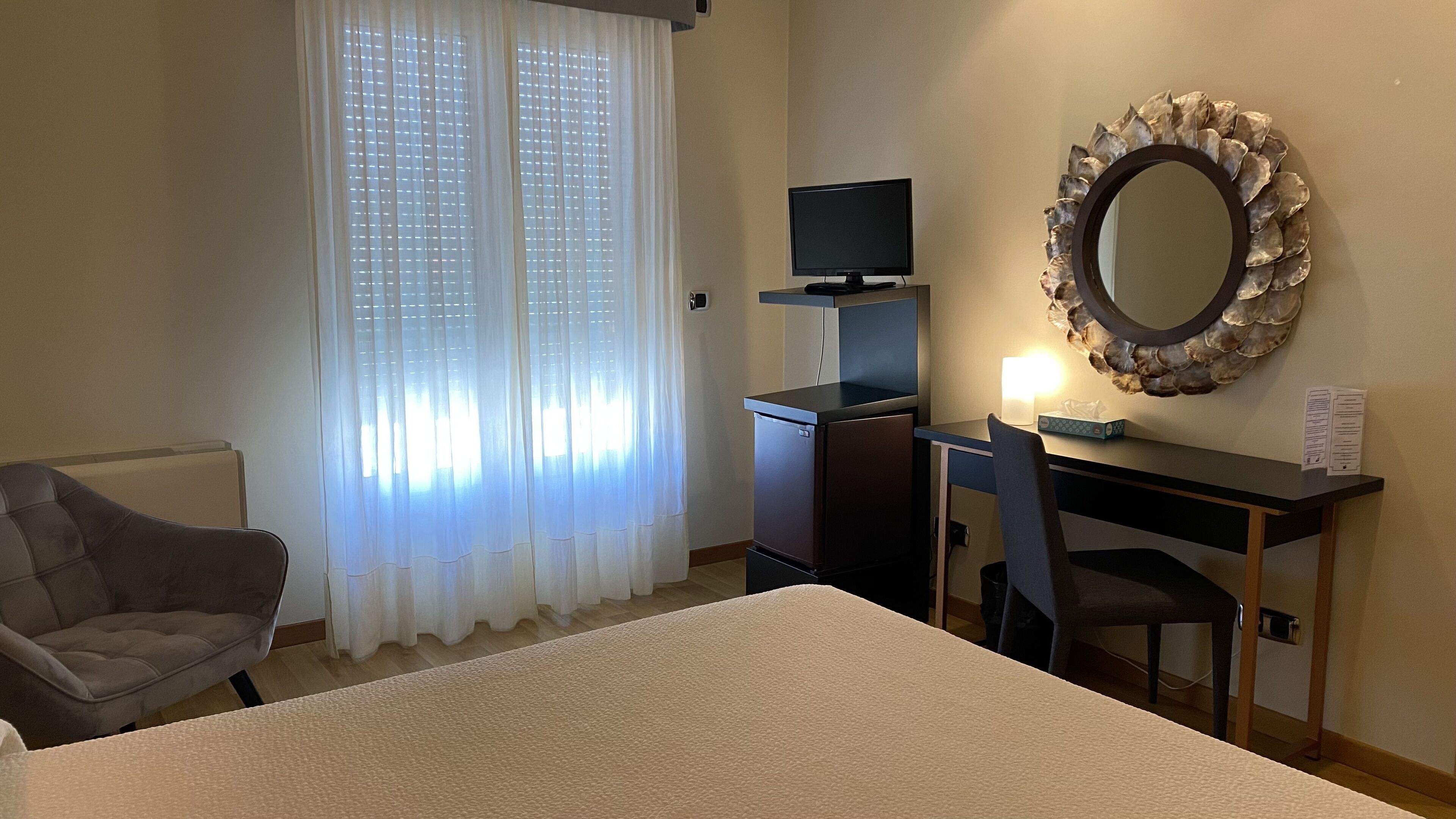A hotel room with a bed, a desk with a computer, a chair, a TV, and a decorative mirror.