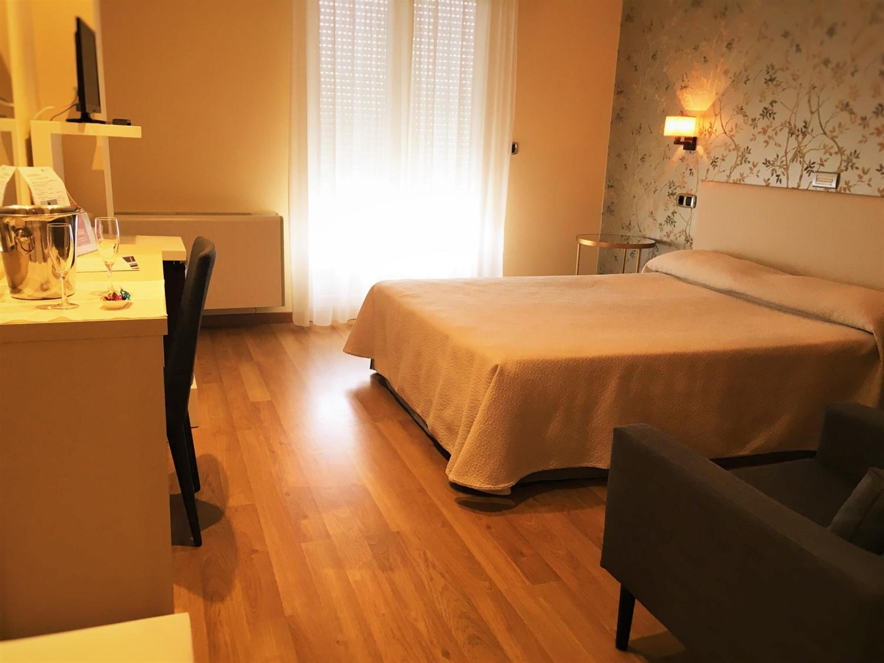 A hotel room with a bed, a desk with a computer, a chair, and a bedside table with a lamp.