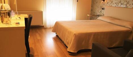 A hotel room with a bed, a desk with a computer, a chair, and a bedside table with a lamp.