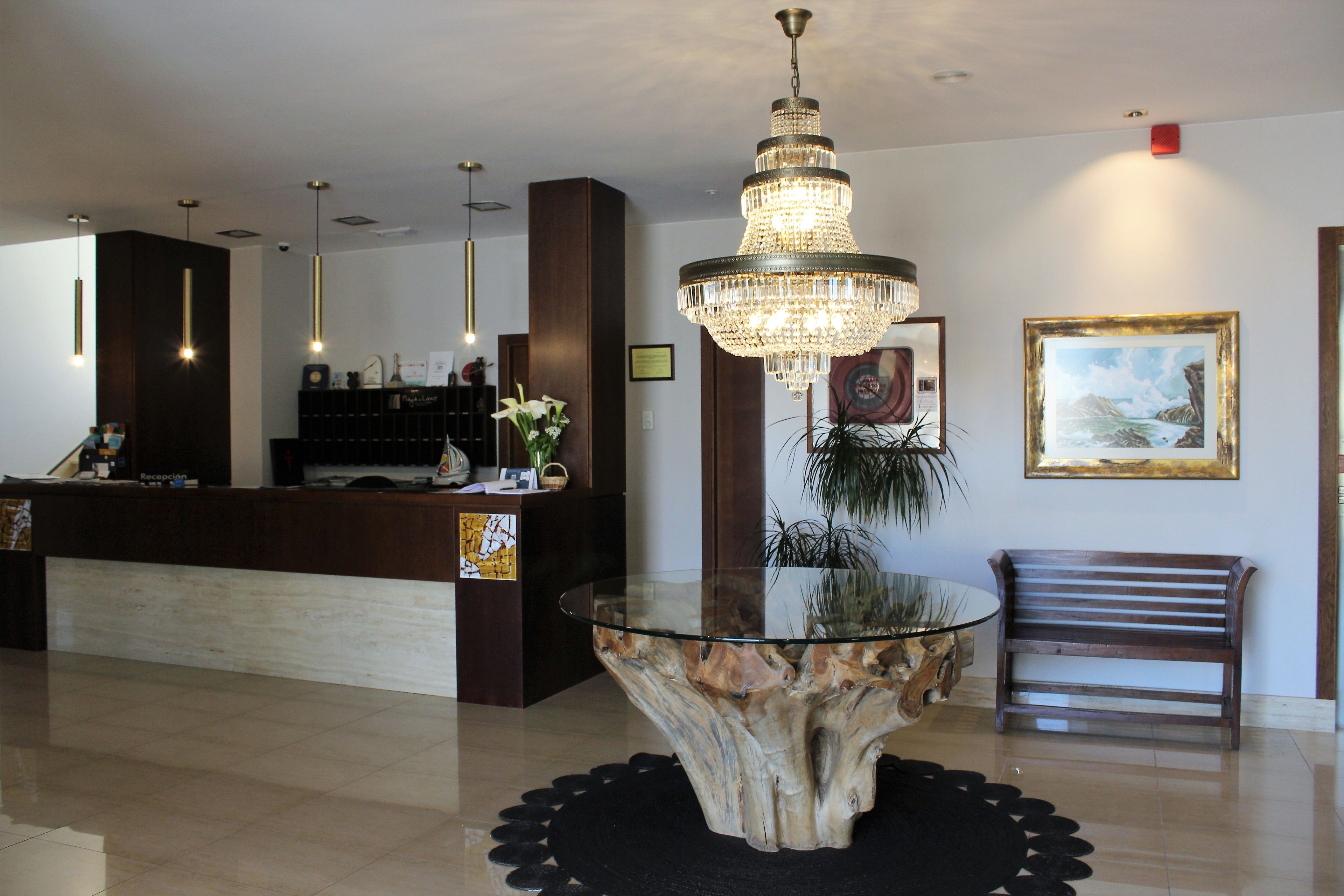 A reception area with a glass-top coffee table, a wooden bench, a chandelier, and a framed painting on the wall.