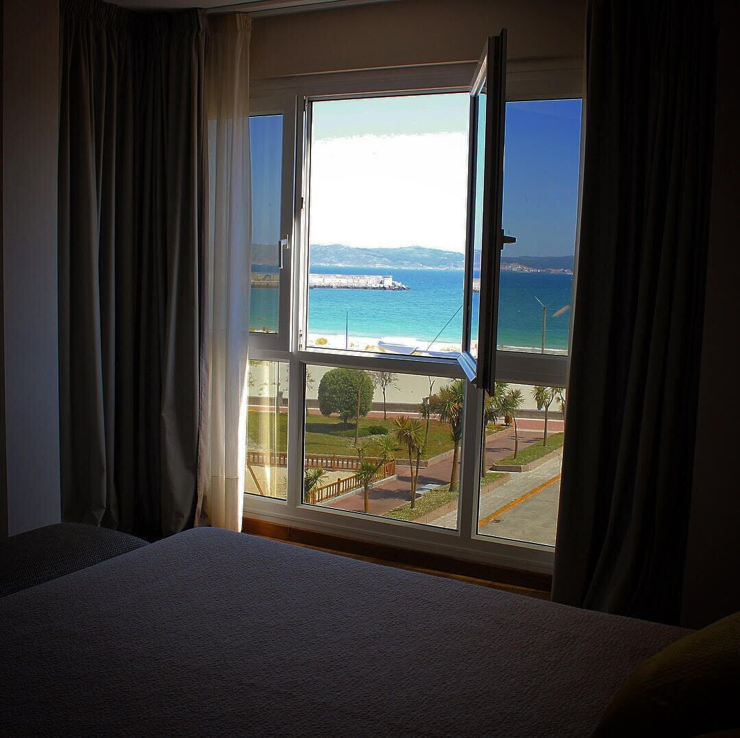 A hotel room with a large window offering a view of a beach, a pier, and a distant mountain range.