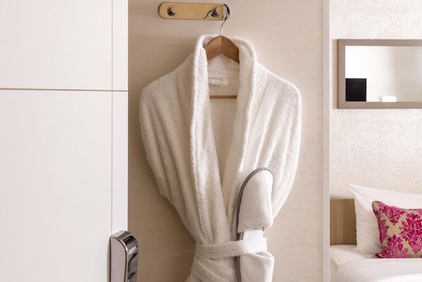 A white robe hanging on a wooden hanger in a hotel room with a wooden headboard, a mirror, and a pink pillow.