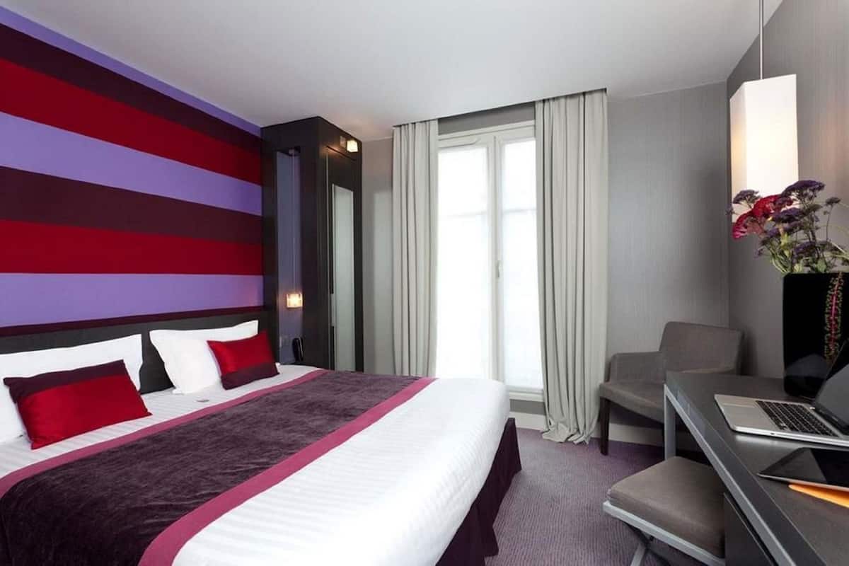 A hotel room with a bed, a desk, a chair, a window with curtains, and a wall with horizontal stripes.