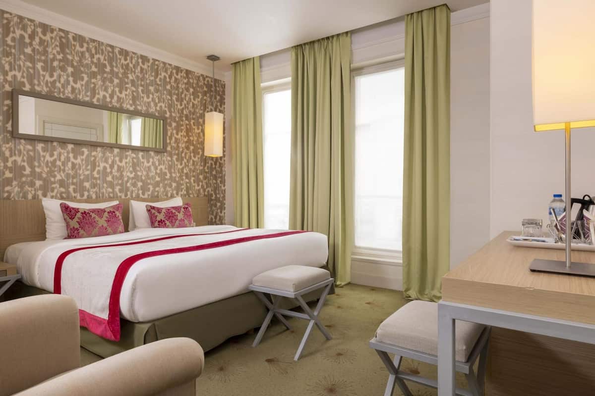 A hotel room with a bed, a desk, a chair, a small stool, a mirror, and a window with curtains.