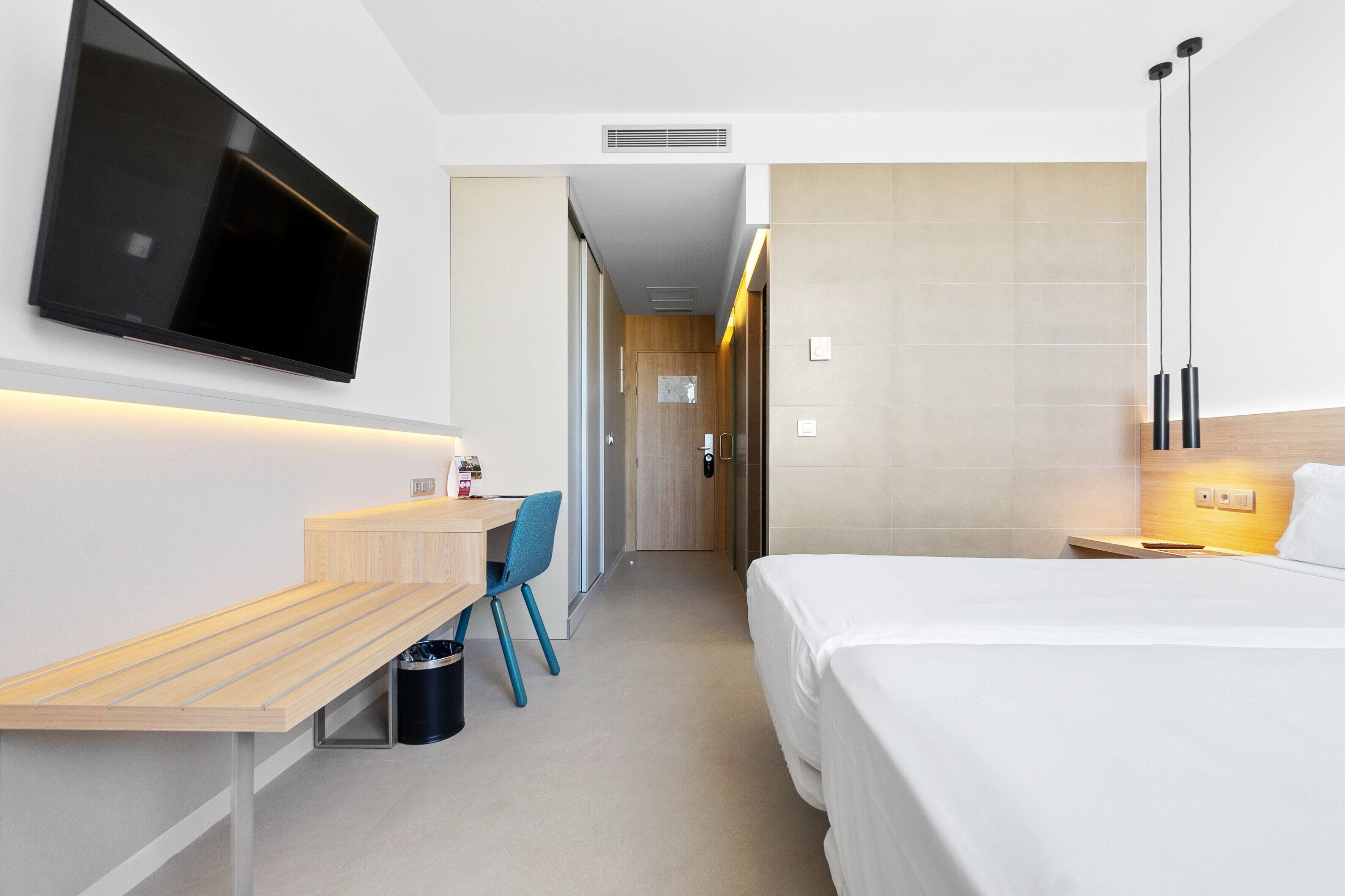 A modern hotel room with a flat-screen TV mounted on the wall, a wooden desk with a chair, a bed with white bedding, and a bedside table with a lamp.