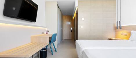 A modern hotel room with a flat-screen TV mounted on the wall, a wooden desk with a chair, a bed with white bedding, and a bedside table with a lamp.