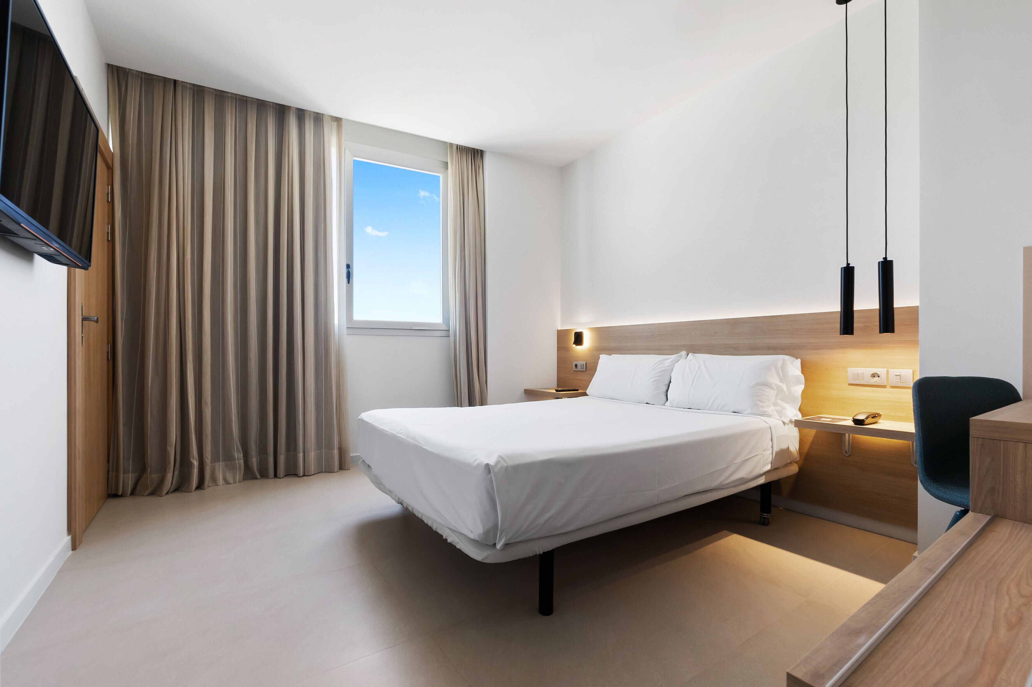 A modern hotel room with a bed, bedside table, lamp, and a desk with a chair.