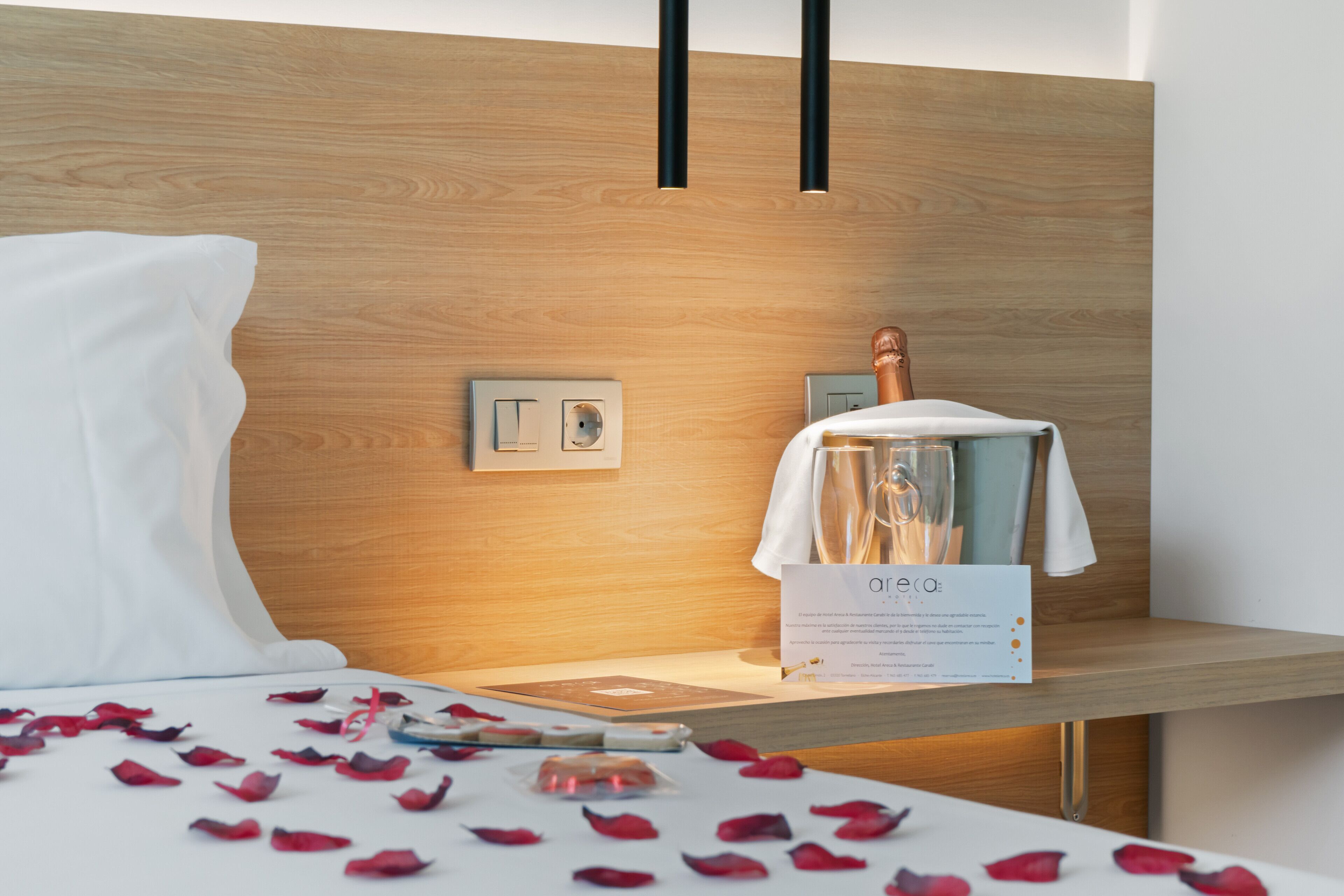 Una habitación de hotel con cabecera de madera, una cama con pétalos de rosa, una copa de champán y un menú en la mesita de noche.