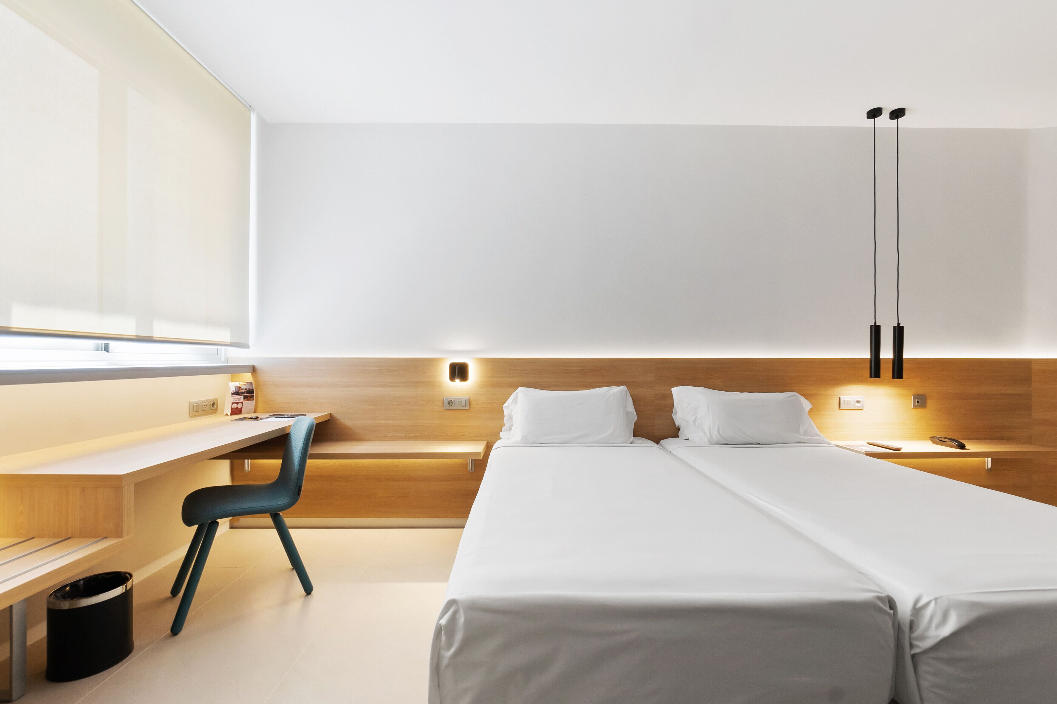 Una habitación de hotel moderna con cabecera de madera, un escritorio con silla, dos camas y un ventanal grande.