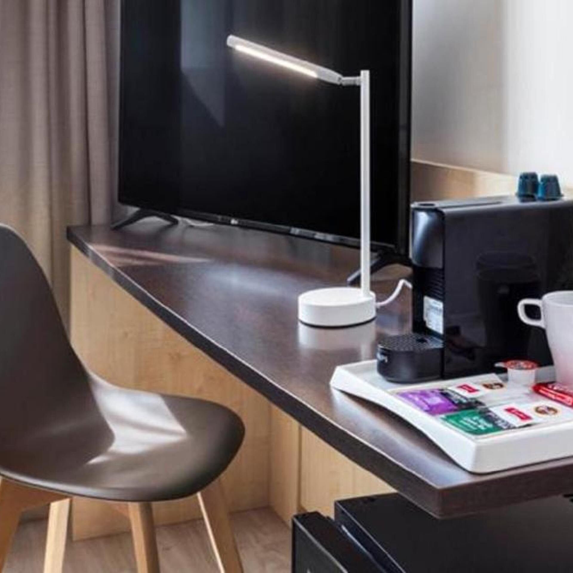 Una habitación de hotel moderna con un escritorio de madera, una silla negra, un televisor de pantalla plana, una lámpara de escritorio, una cafetera y una bandeja con cápsulas de café y una revista.