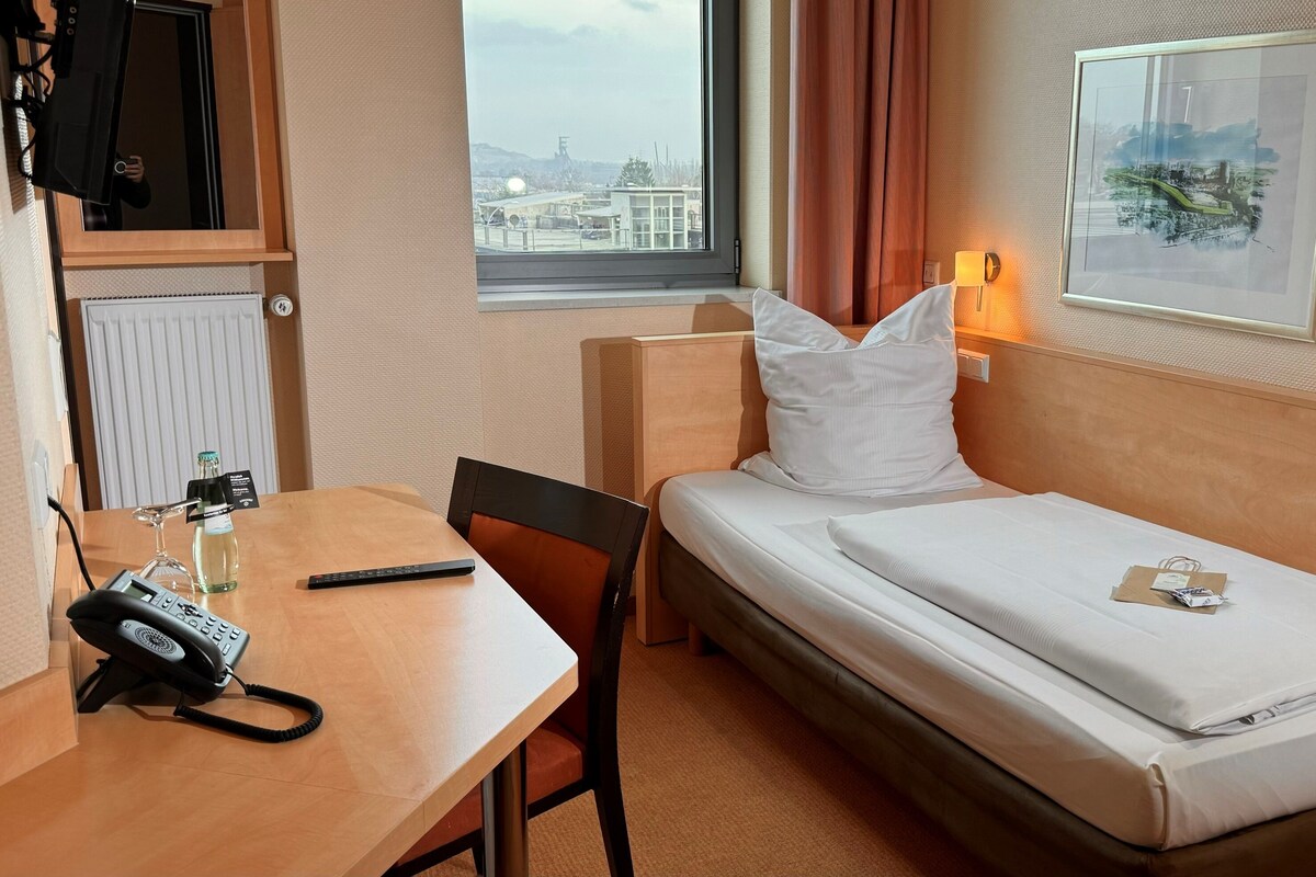 A hotel room with a bed, a desk with a telephone, a chair, a window with a view of buildings, and a framed picture on the wall.