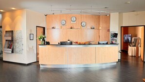 A hotel reception area with a wooden reception desk, three wall clocks, a map, and a flower vase. - Hotel Rhein-Ruhr Bottrop Hauptbahnhof by Mayburgh (Bottrop)
