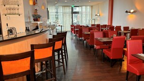 A restaurant interior with wooden bar counters, red upholstered chairs, and a long counter with a glass display of glasses. - Hotel Rhein-Ruhr Bottrop Hauptbahnhof by Mayburgh (Bottrop)