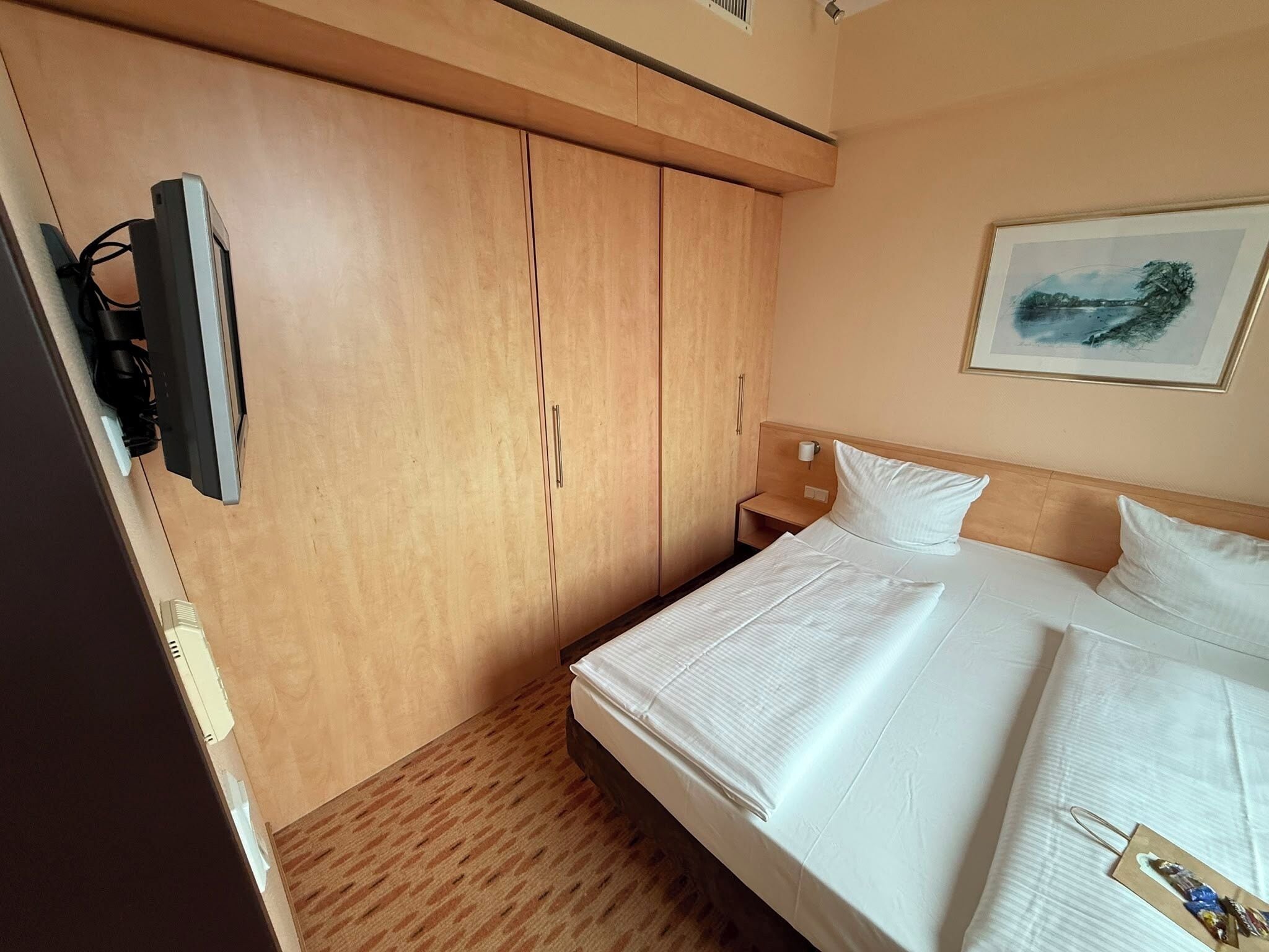Habitación de hotel con cama, televisor montado en la pared, mesita de noche y un cuadro.