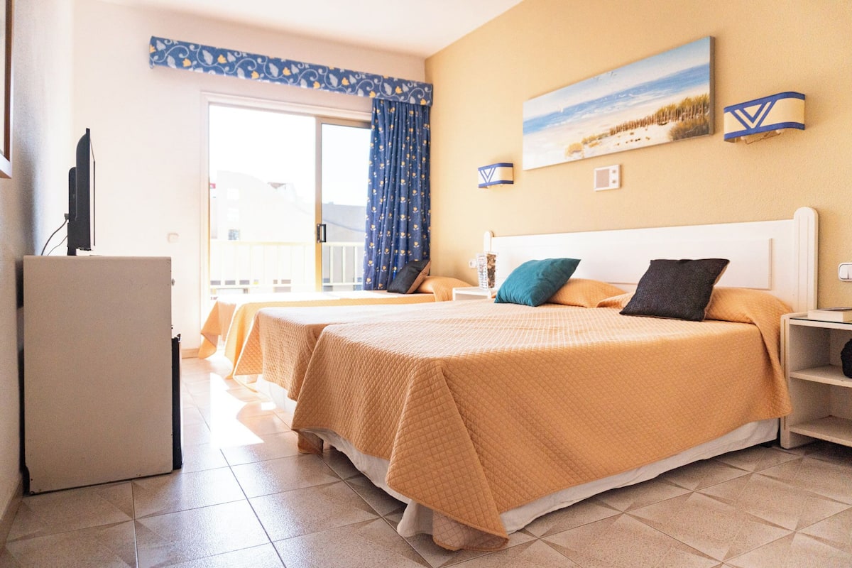 Ein Hotelzimmer mit zwei Betten, einem Fernseher, einem Balkon und einem Gemälde mit einer Strandansicht.