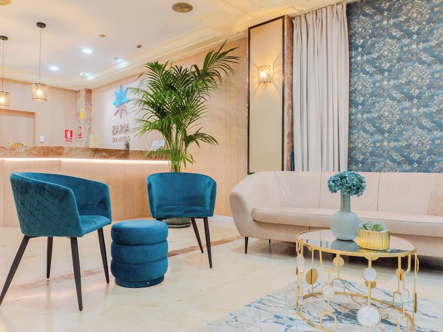 A hotel reception area with a marble reception desk, teal chairs, a beige sofa, and a decorative table with a vase.