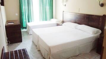 Habitación de hotel con dos camas individuales, cabecera de madera, mesita de noche pequeña, cesto de basura y una ventana con cortinas verdes.