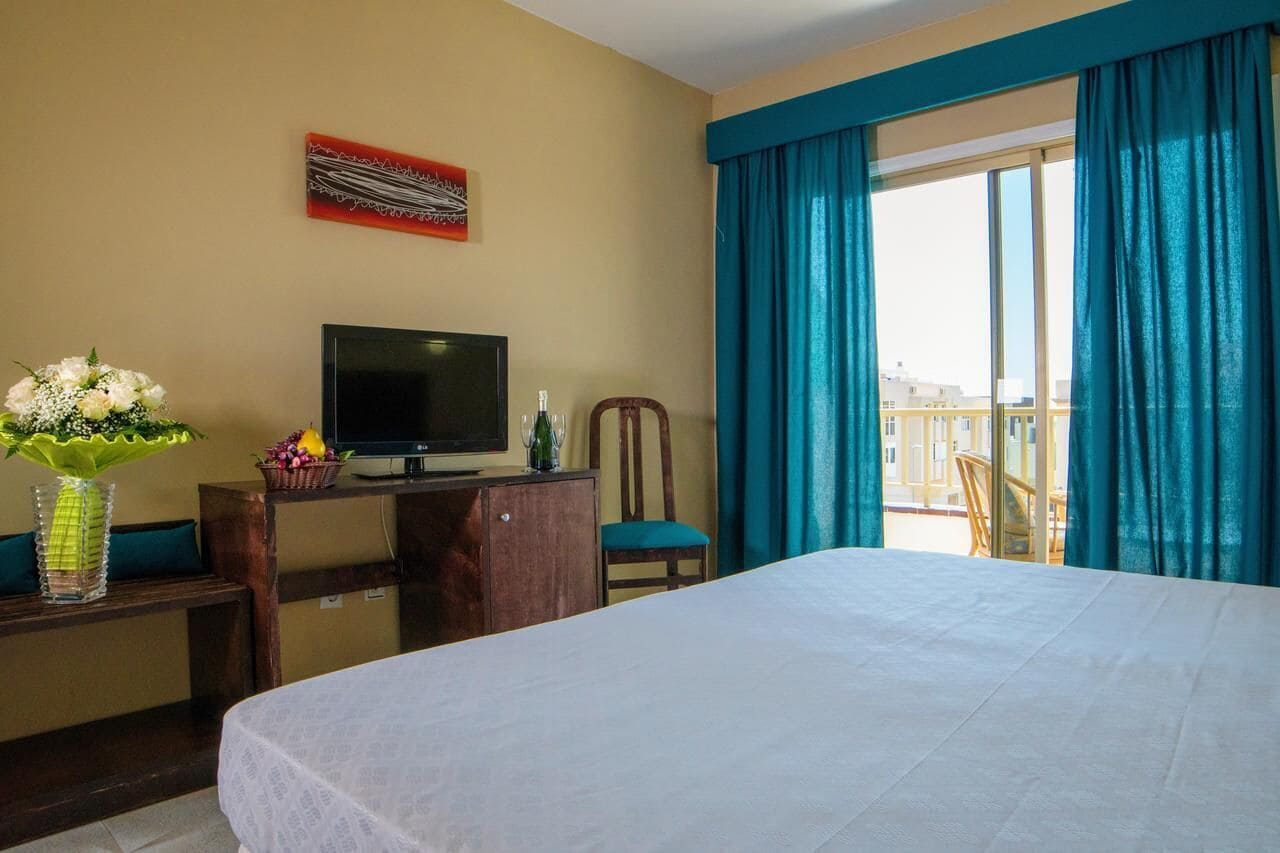 A hotel room with a bed, a desk with a TV, a chair, a vase with flowers, and a balcony with curtains.