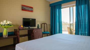 A hotel room with a bed, a desk with a TV, a chair, a vase with flowers, and a balcony with curtains.