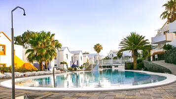 Zona de piscina con sillones de descanso y palmeras, rodeada de edificios con balcones.