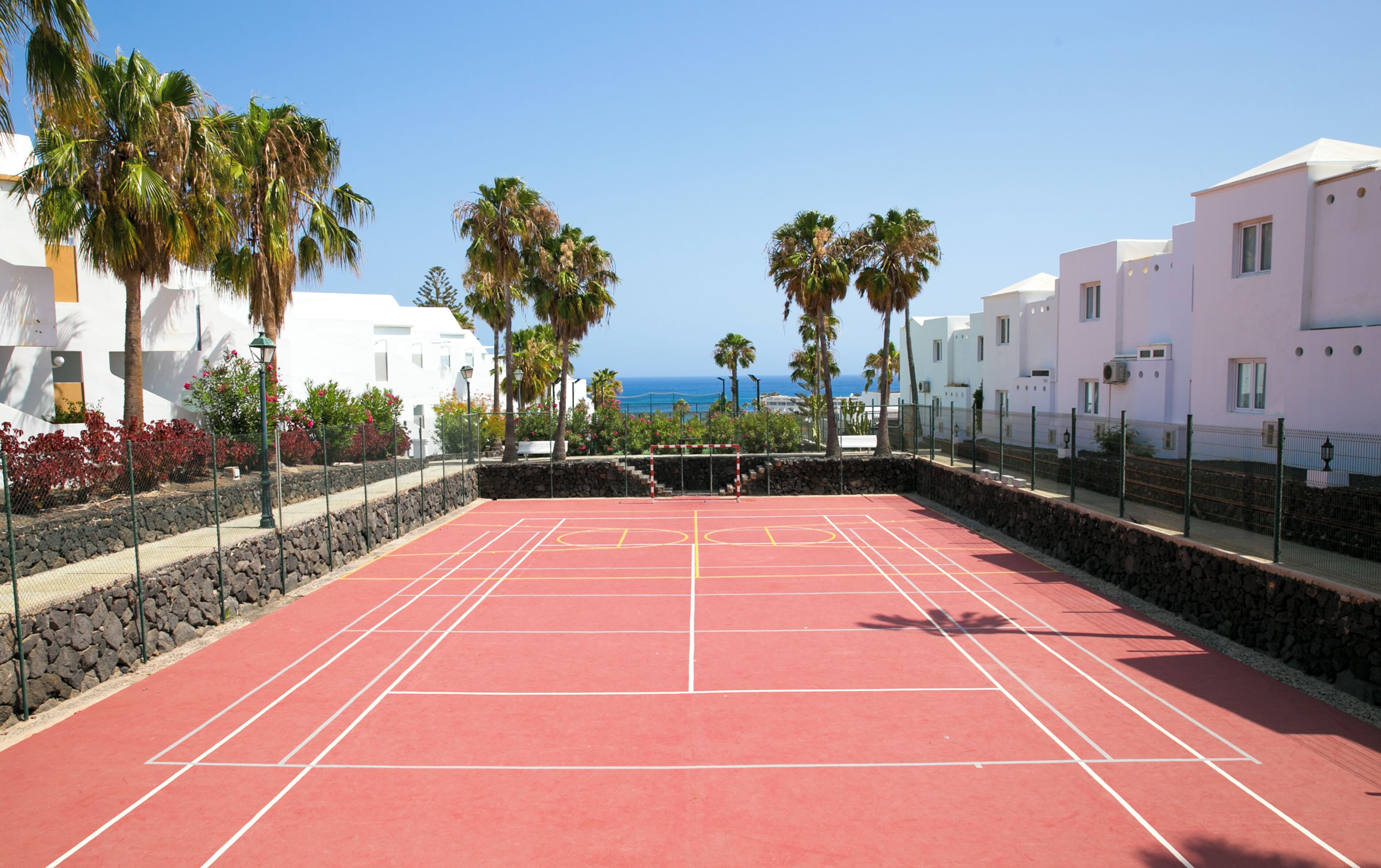Un court de tennis avec un filet, entouré de murs de pierre et de palmiers, avec des bâtiments en arrière-plan.
