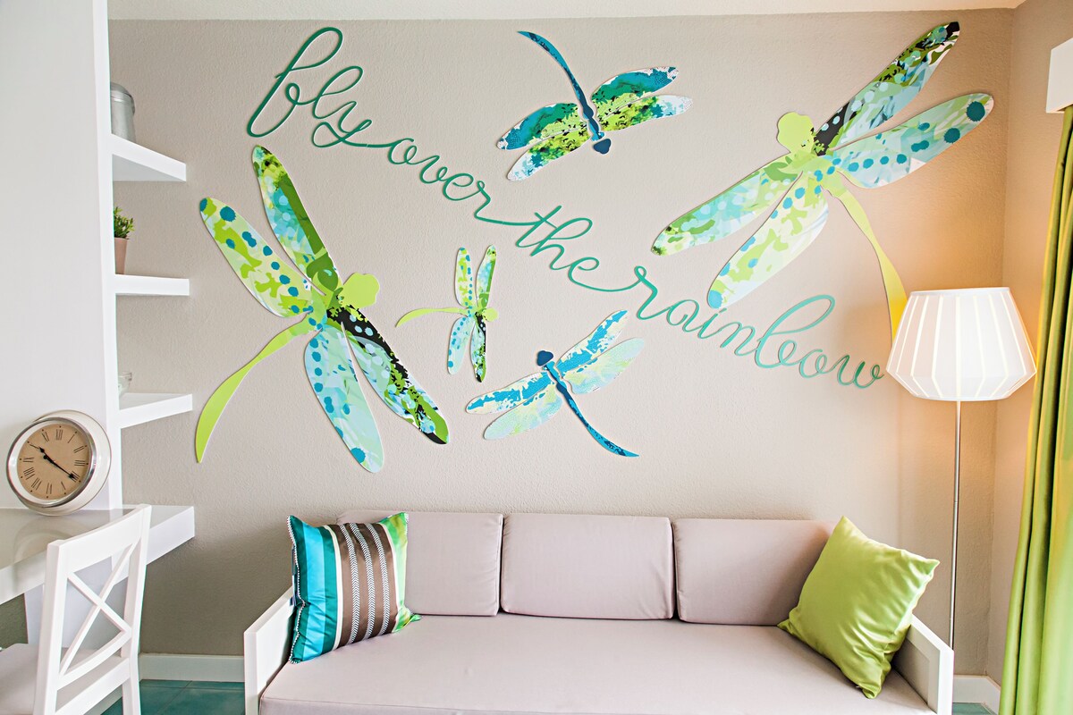 A room with a beige sofa, a clock, and wall art featuring dragonflies and the phrase "fly over the rainbow".