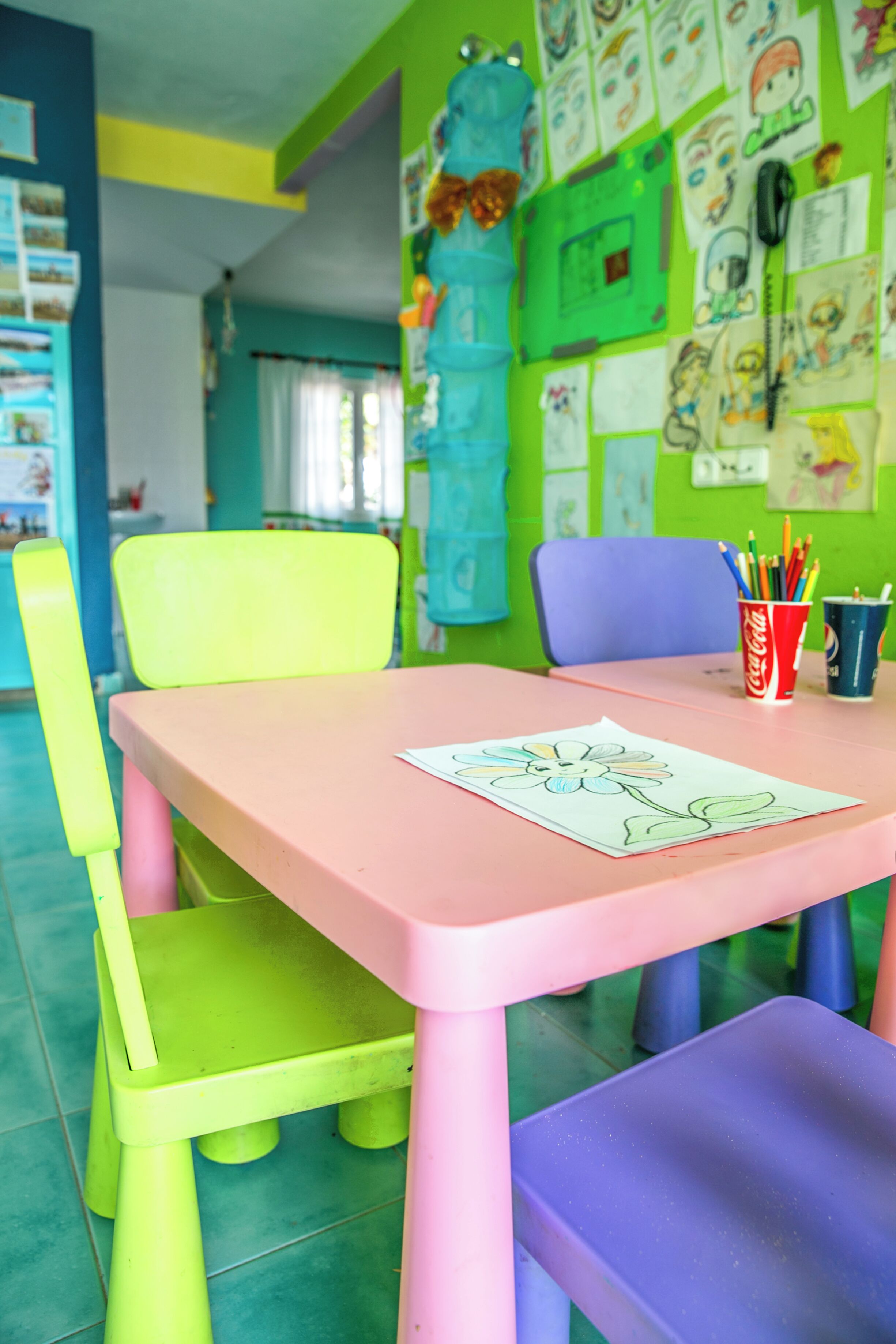 Ein helles Zimmer mit einem rosafarbenen Tisch, grünen und violetten Stühlen sowie einer Wand, die mit Kinderzeichnungen und Notizen verziert ist.