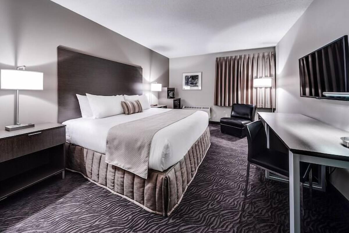 A hotel room with a bed, bedside table, lamp, chair, desk, and a large mirror.