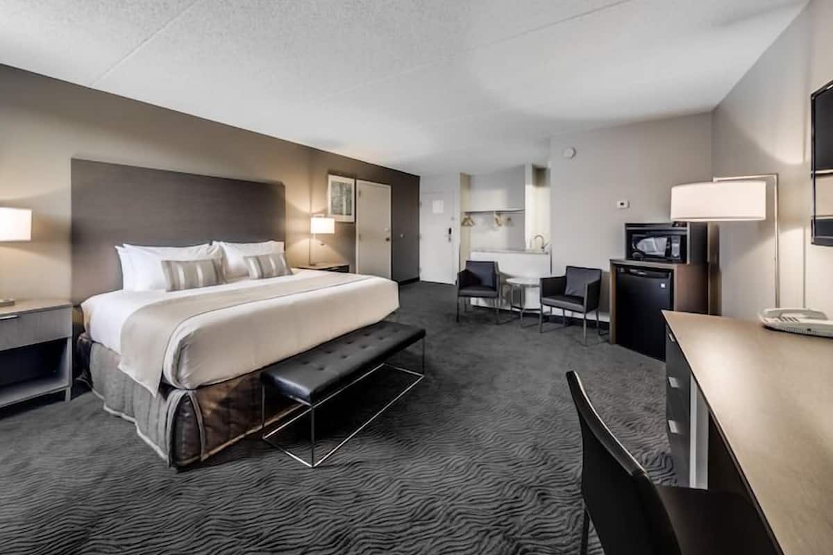 A hotel room with a large bed, a desk, a chair, a microwave, a small table, and a bench.