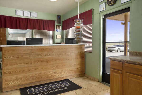 A reception desk with a wooden finish, a ViewSonic monitor, and an Acer monitor. There is a window with blinds and a red curtain. A doormat with "Travelodge Holbrook, AZ" is visible. - Travelodge by Wyndham Holbrook (Holbrook)