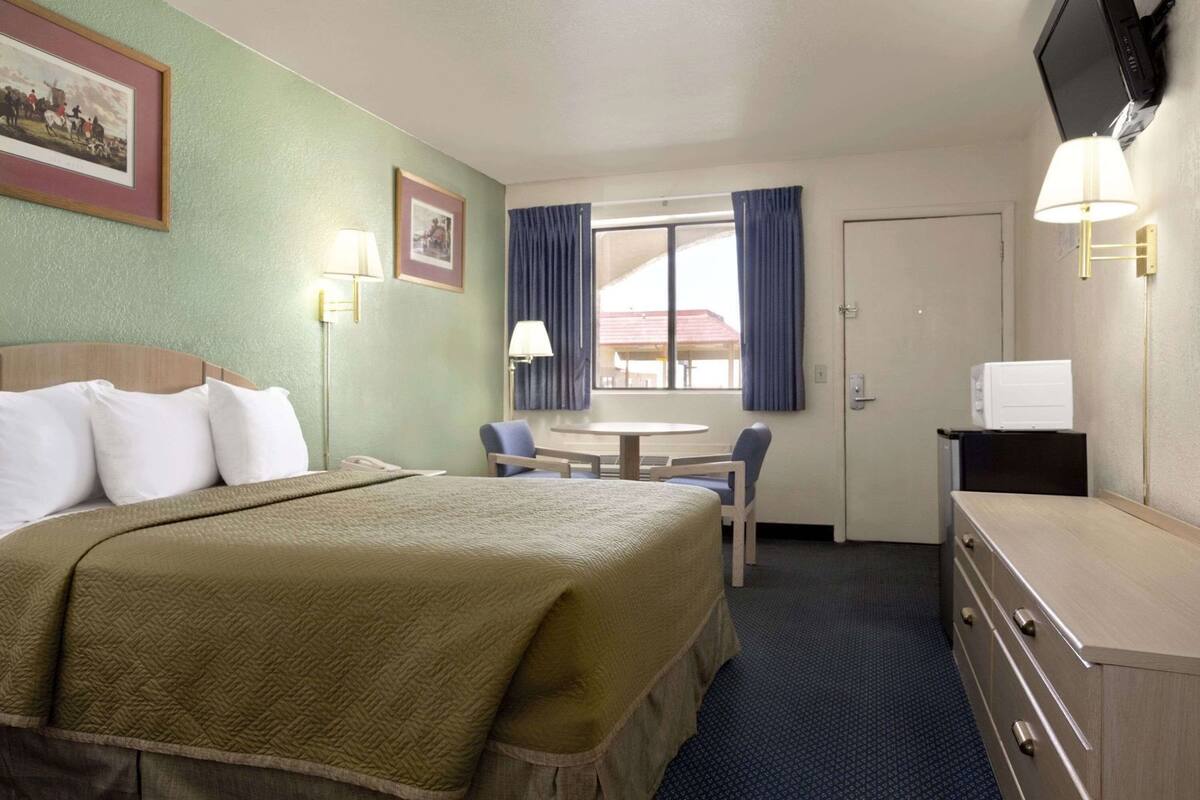 Une chambre d’hôtel avec un lit, un bureau, deux chaises, une télévision et une fenêtre avec des rideaux.