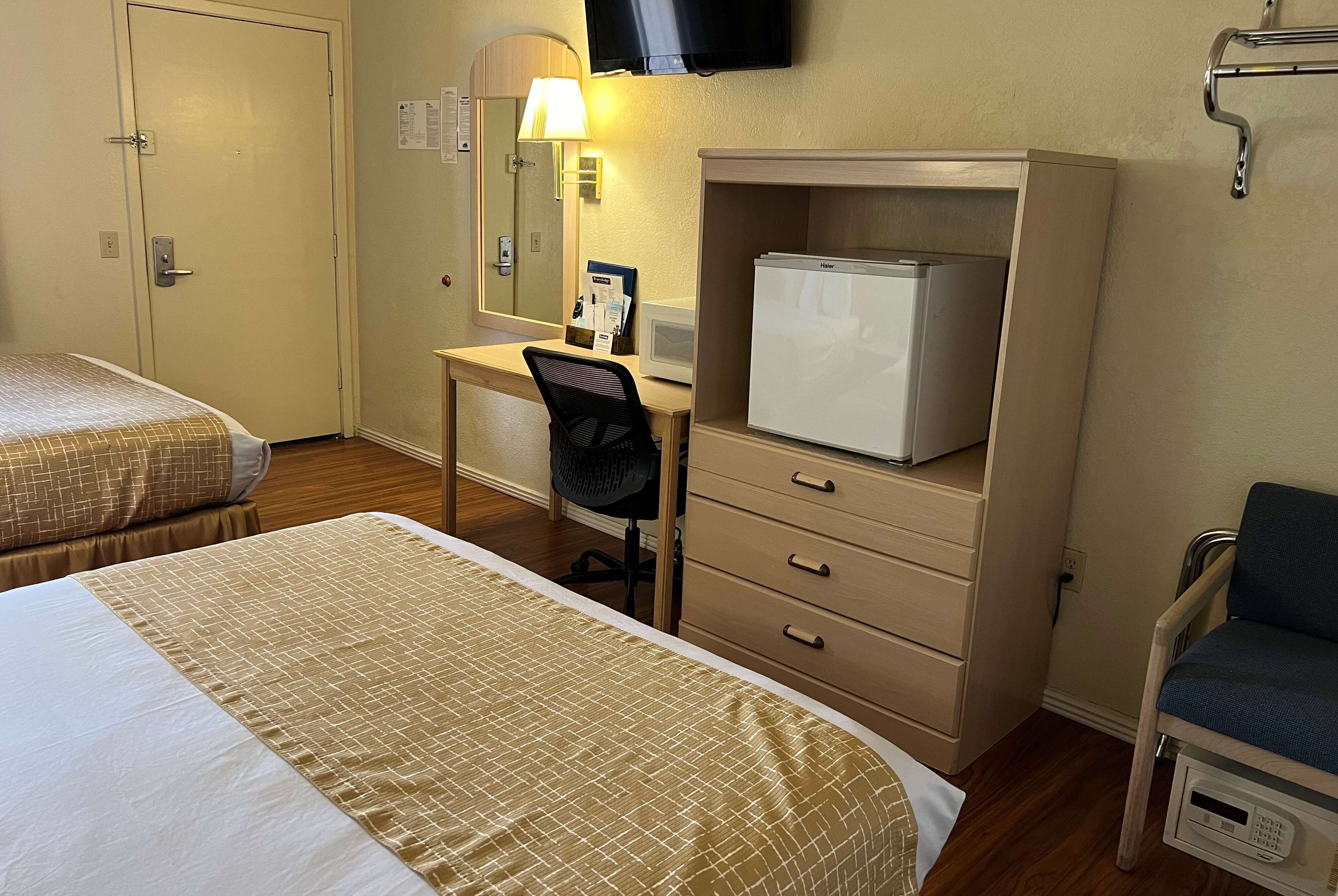 A hotel room with two beds, a desk with a chair, a refrigerator, a TV, and a chair with a tray table.