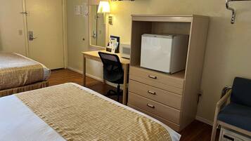 A hotel room with two beds, a desk with a chair, a refrigerator, a TV, and a chair with a tray table.