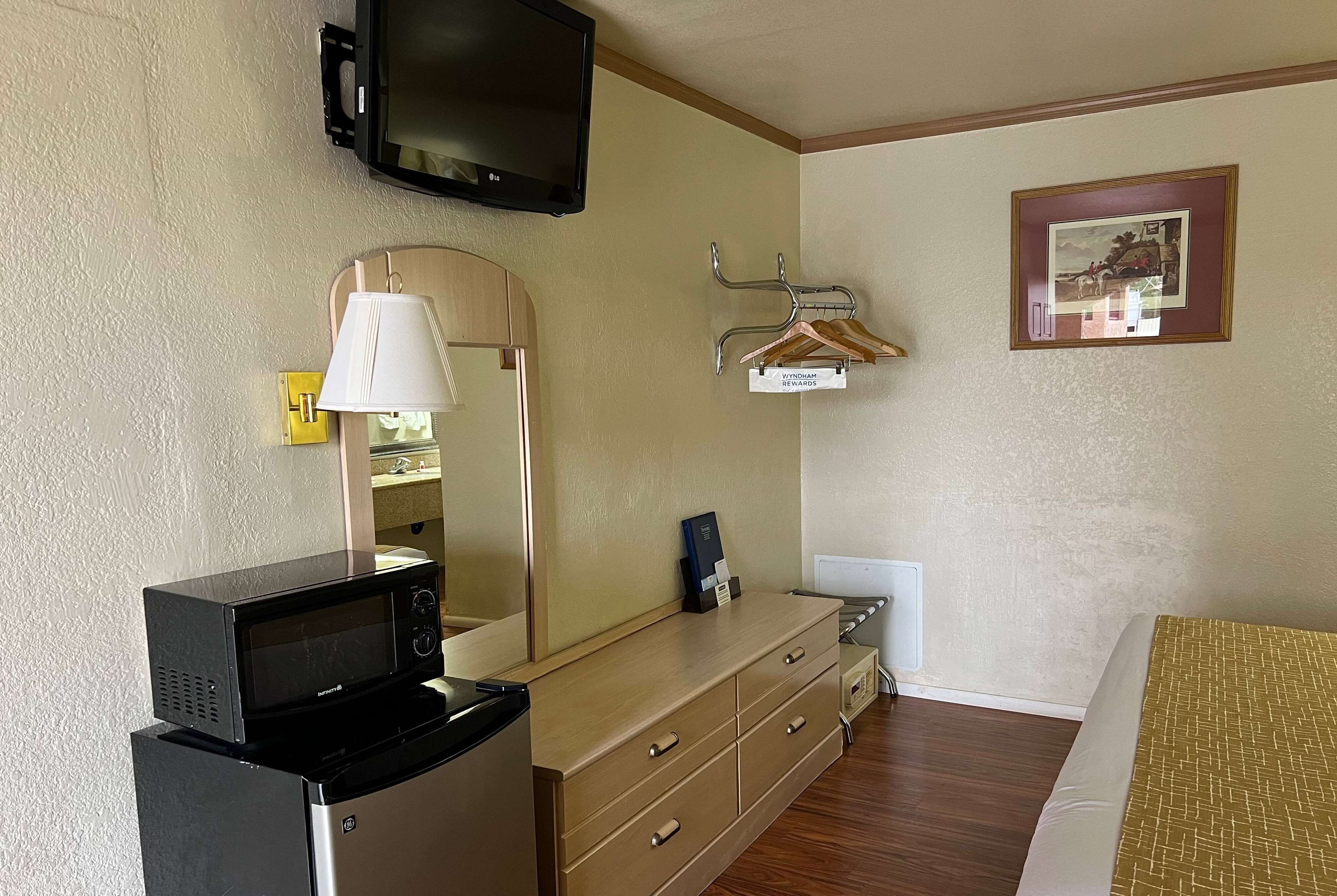 Habitación de hotel con microondas sobre un refrigerador pequeño, un televisor montado en la pared, una cómoda con cajones, un perchero y un cuadro enmarcado en la pared.