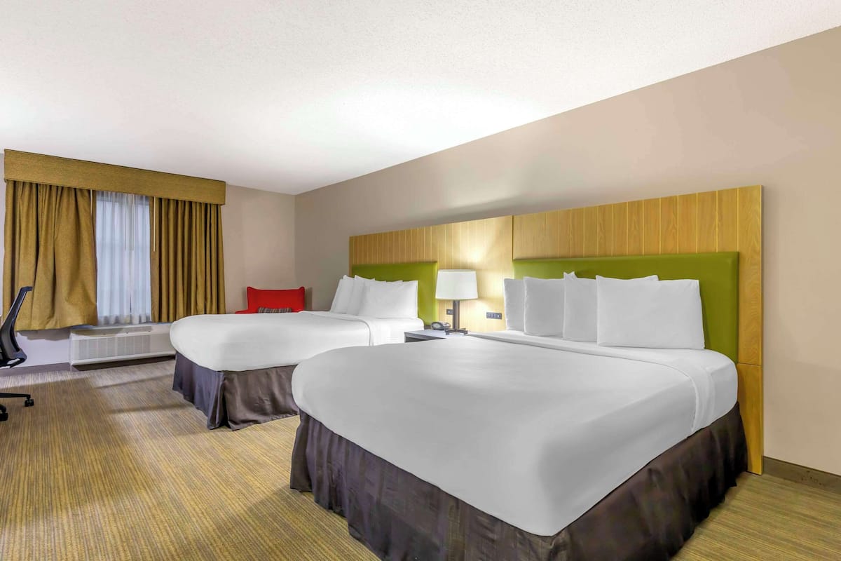Una habitación de hotel con dos camas, un almohadón rojo, una lámpara y una ventana con cortinas.