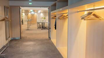 Pasillo de hotel con piso alfombrado, un área de descanso con mesas y sillas, y un espacio para guardar ropa con perchas.