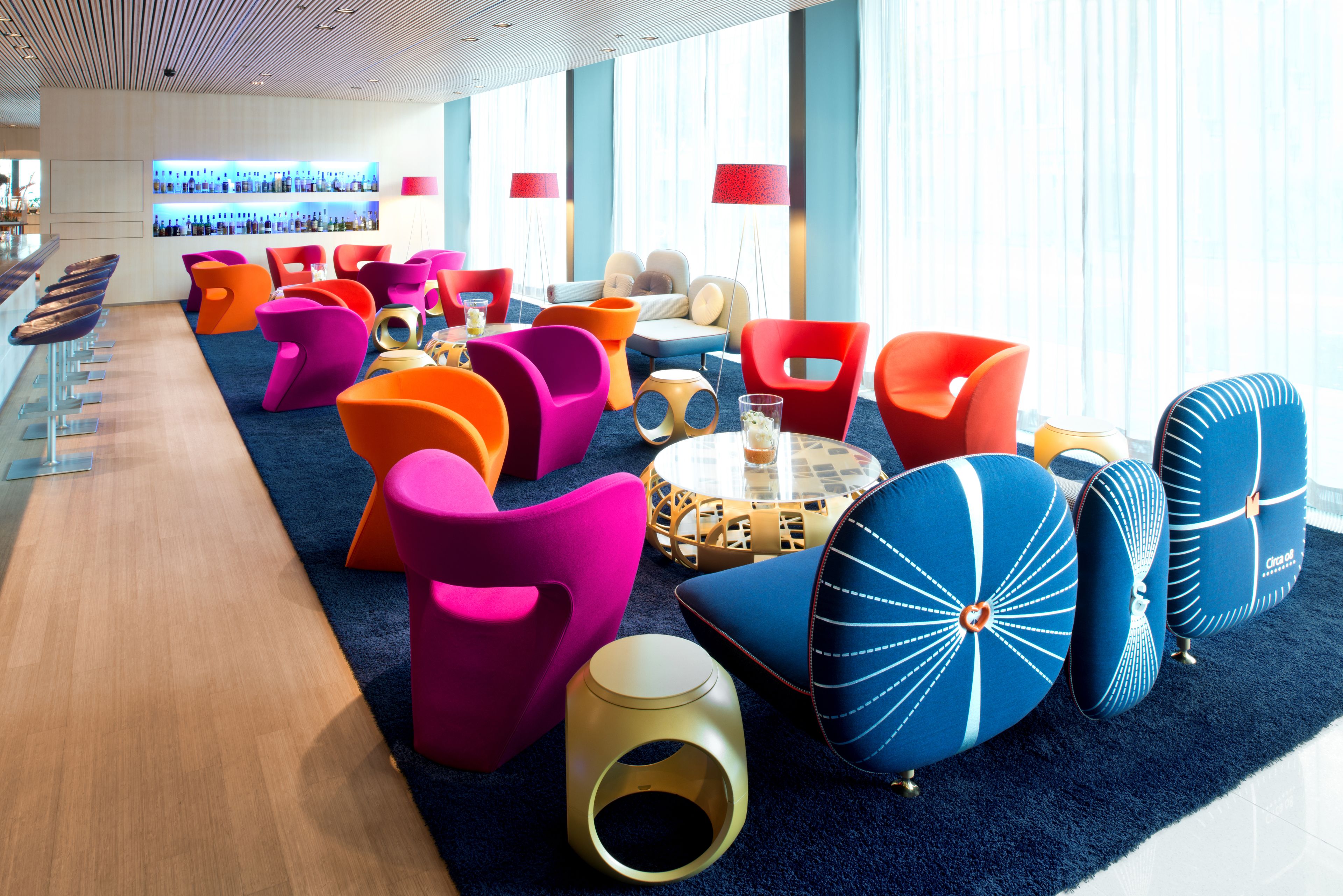 A modern lounge area with vibrant seating options, a bar, and a view of the city through large windows.