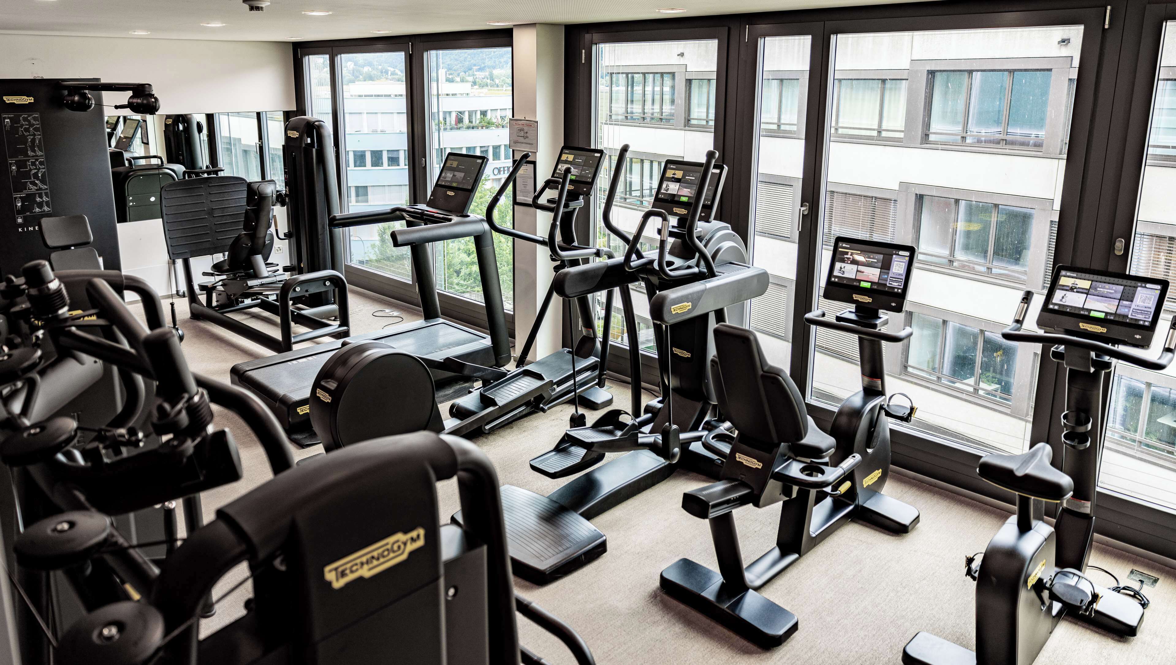 A gym with various exercise machines, large windows, and a view of buildings outside.