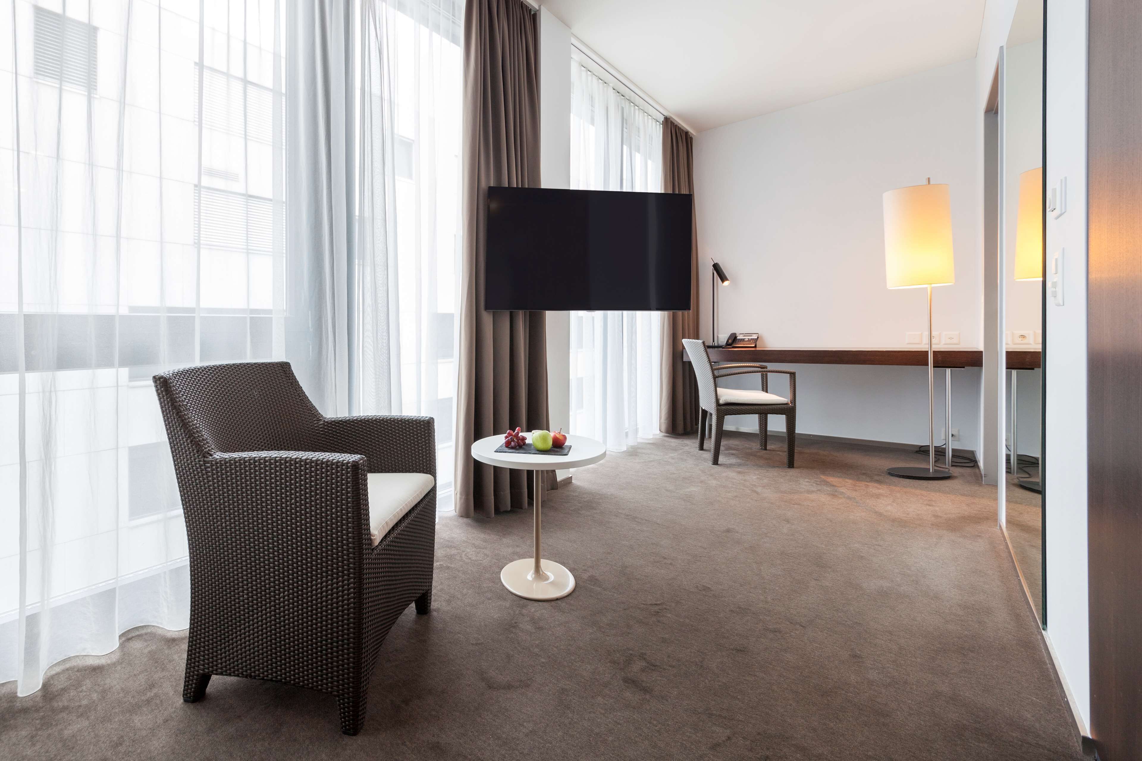 Habitación de hotel con un ventanal grande, un televisor de pantalla plana, una mesa redonda con fruta, una silla de mimbre y un escritorio con lámpara.