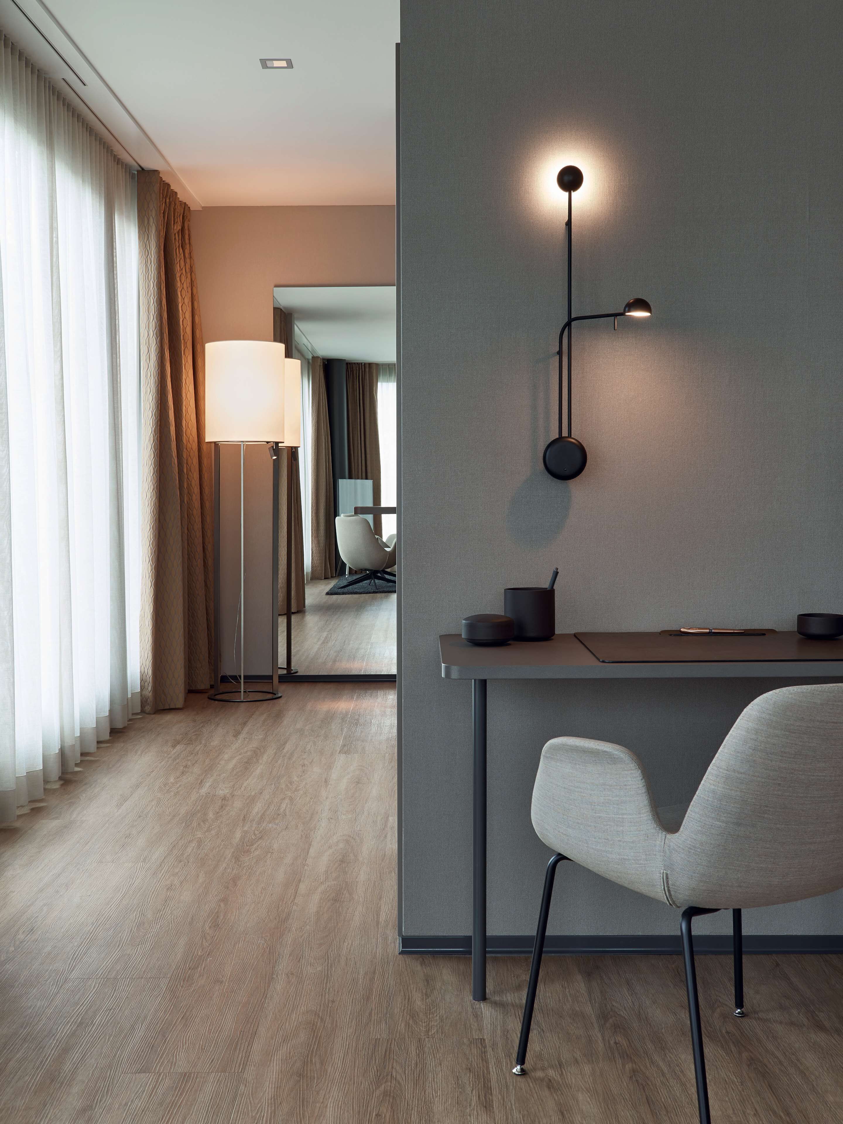 A modern hotel room with a wooden floor, a desk with a lamp, a chair, and a wall-mounted light fixture.
