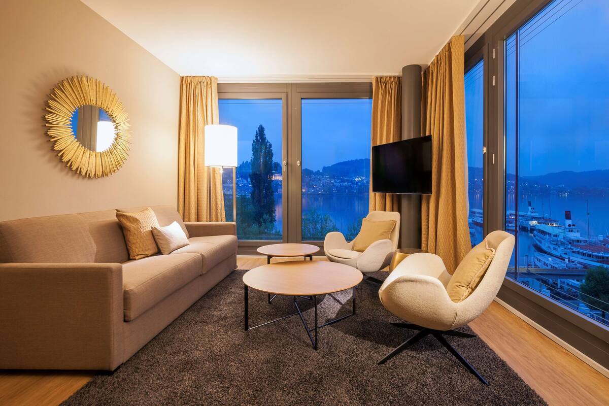 A modern hotel room with a sofa, two armchairs, a coffee table, a TV, and a large window with a city view.