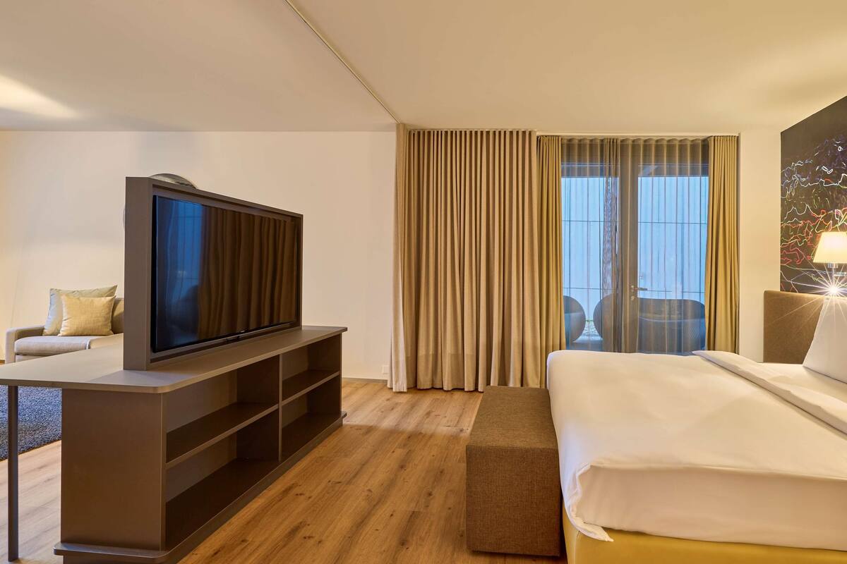 A hotel room with a flat-screen TV, a sofa, a bed, and a large window with curtains.