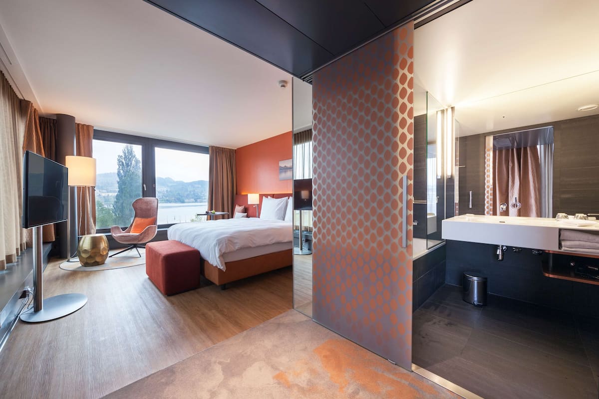 A modern hotel room with a bed, a seating area with a chair, a TV, and a bathroom with a mirror and sink.