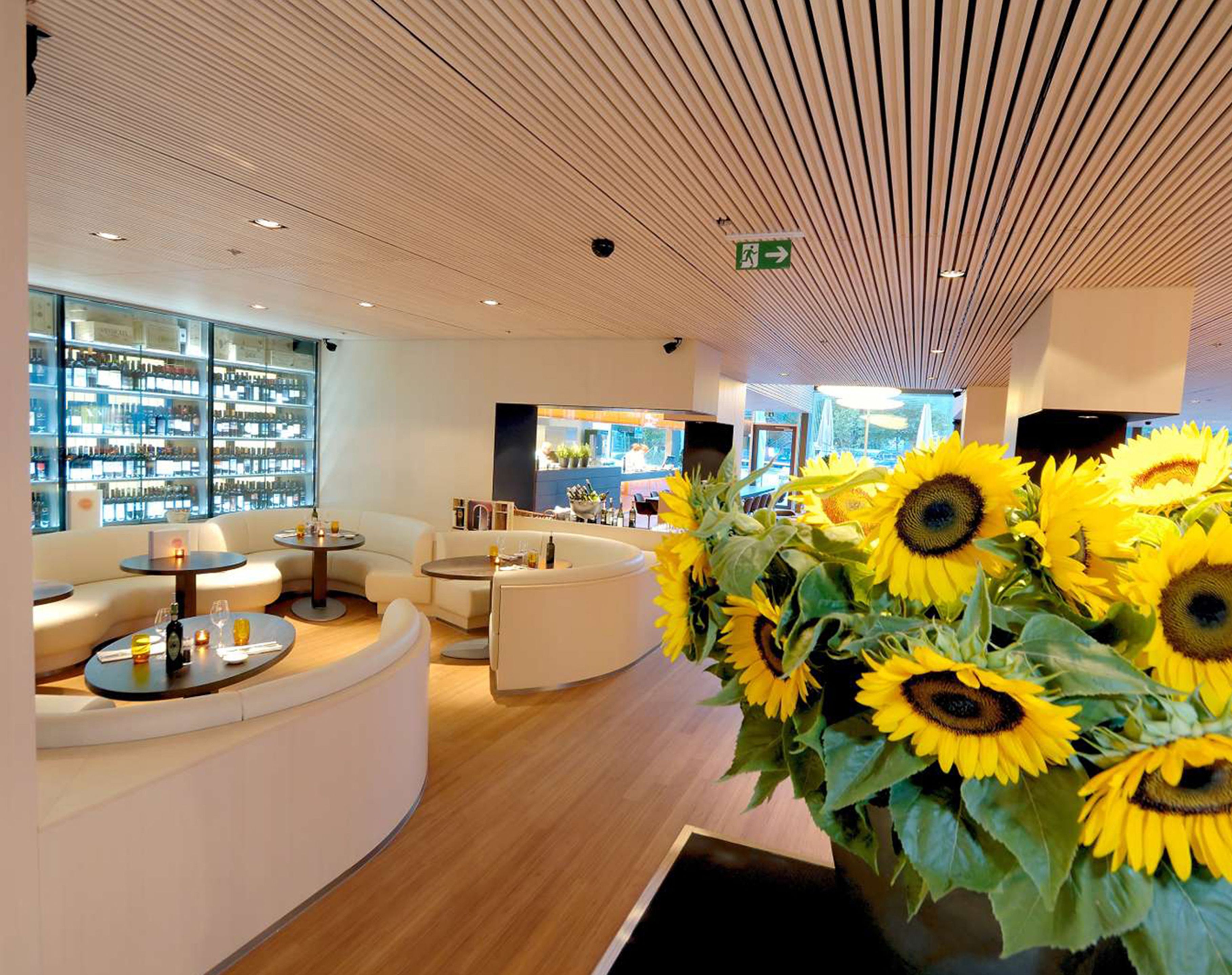 A modern interior with a curved reception desk, seating areas, and a large window displaying a bar area. Sunflowers are prominently displayed in the foreground.
